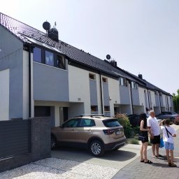 Budownictwo - Szereg identycznych, nowo wybudowanych domów murowanych z grafitową dachówką i antenami satelitarnymi, z zaparkowanym brązowym SUV-em i spacerującą rodziną na brukowanym podjeździe.