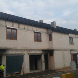 Budownictwo - Budowa szeregowca z białego bloczka, widoczne okna z brązowymi ramami, czarny dach, rusztowanie przy ścianie i pracownik w jaskrawej kurtce.