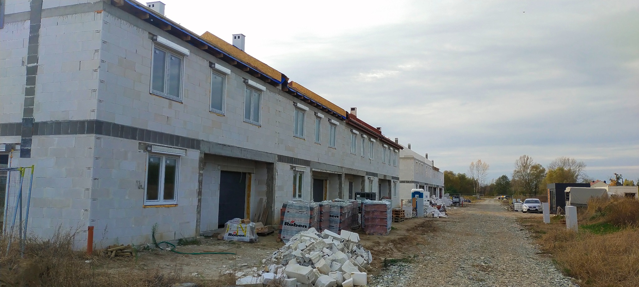 Szeregówka w stanie surowym zamkniętym, z widocznymi bloczkami z betonu komórkowego, częściowo z zamontowanymi roletami zewnętrznymi, z paletami cegieł i gruzem przed budynkiem.