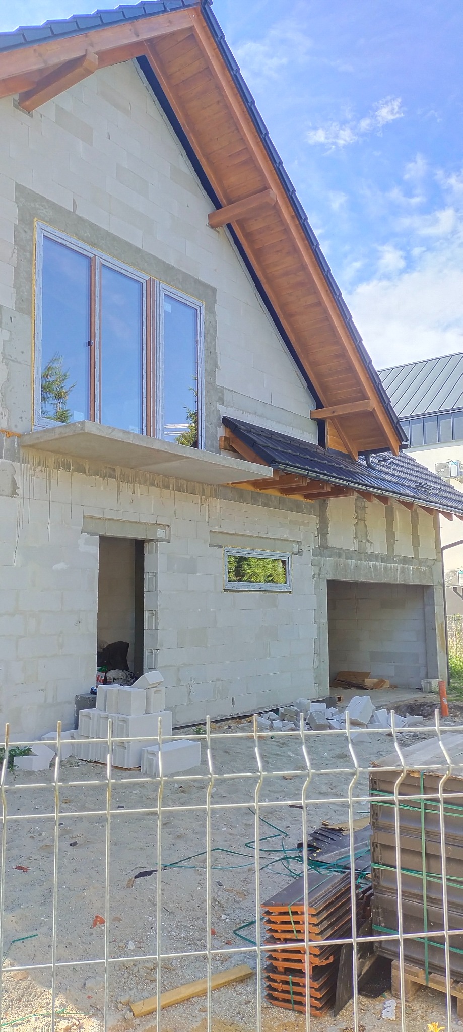 Surowy stan budynku z białych bloczków, widoczne okna z refleksami drzew, drewniana konstrukcja dachu, teren budowy z paletami dachówek i metalowym ogrodzeniem.