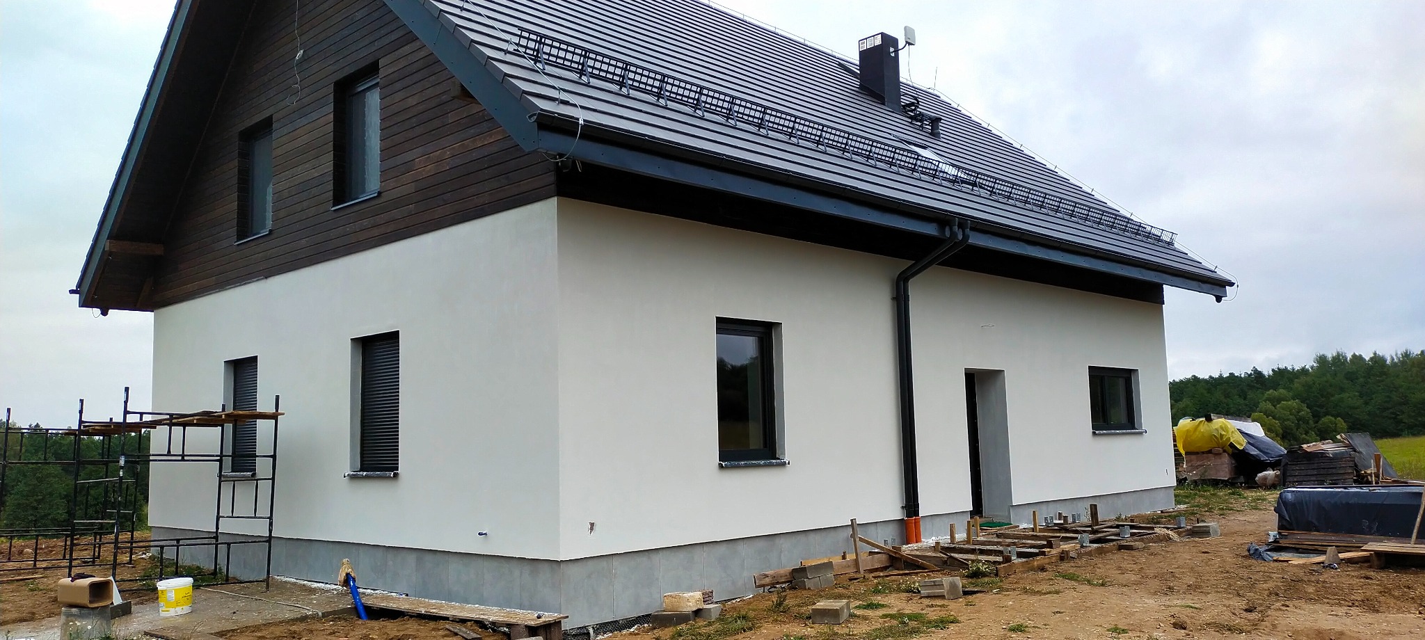 Nowoczesny dom jednorodzinny z ciemnobrązowym wykończeniem pod dachem i czarną dachówką, widoczne rusztowanie z boku i przygotowane szalunki fundamentów, w tle zalesiony teren.