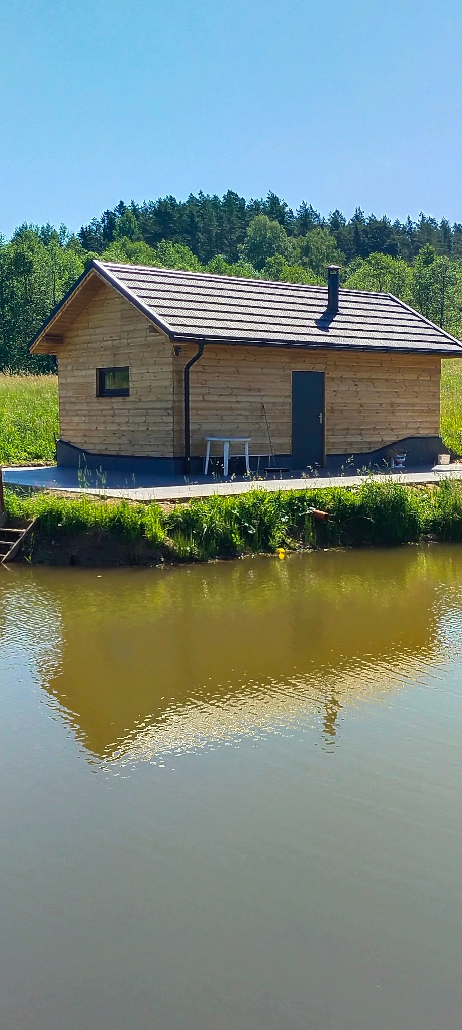 Drewniany domek letniskowy z ciemną stolarką okienną i kominem, posadowiony nad brzegiem stawu, otoczony zielenią.
