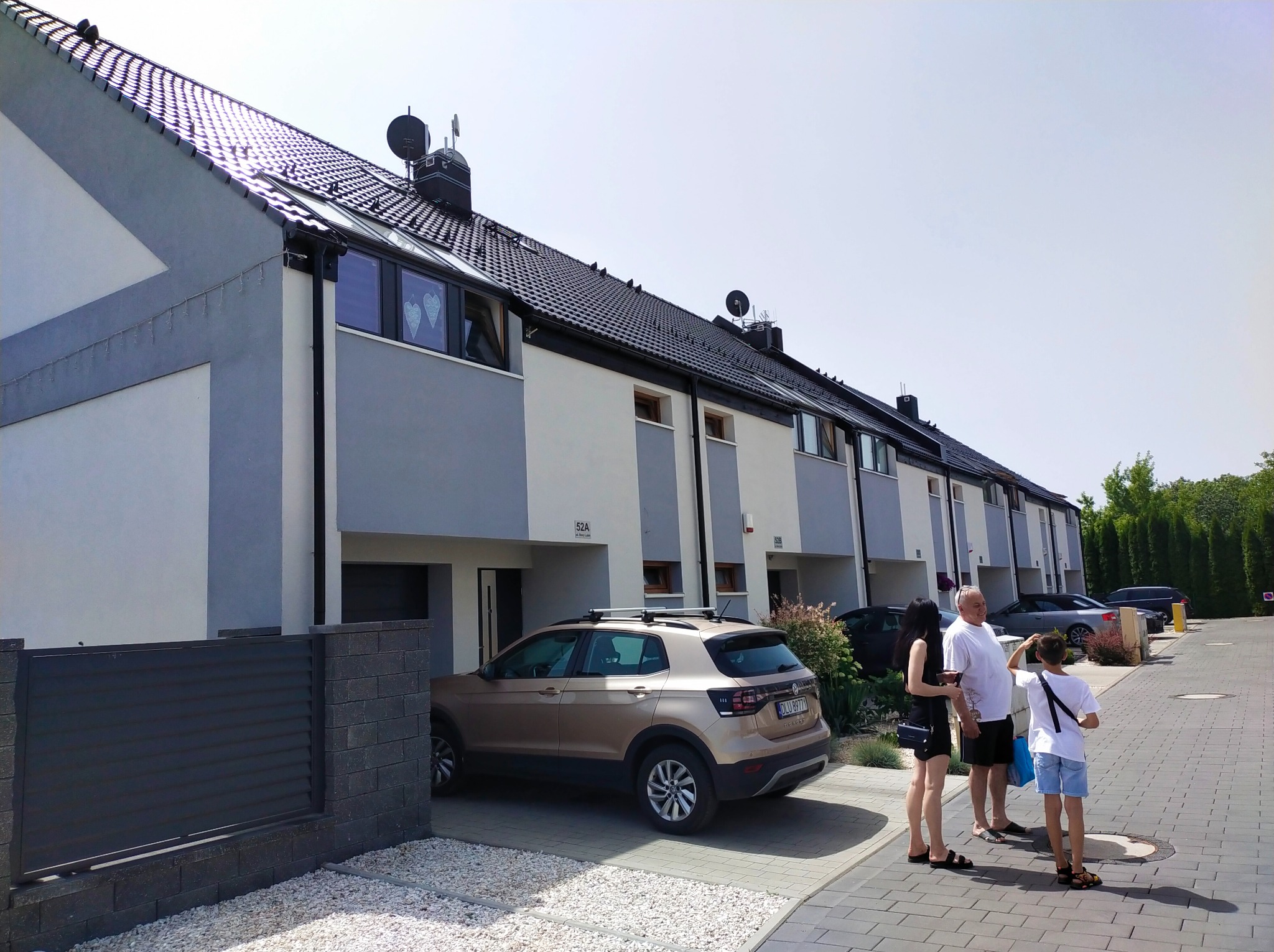 Szereg identycznych, nowo wybudowanych domów murowanych z grafitową dachówką i antenami satelitarnymi, z zaparkowanym brązowym SUV-em i spacerującą rodziną na brukowanym podjeździe.