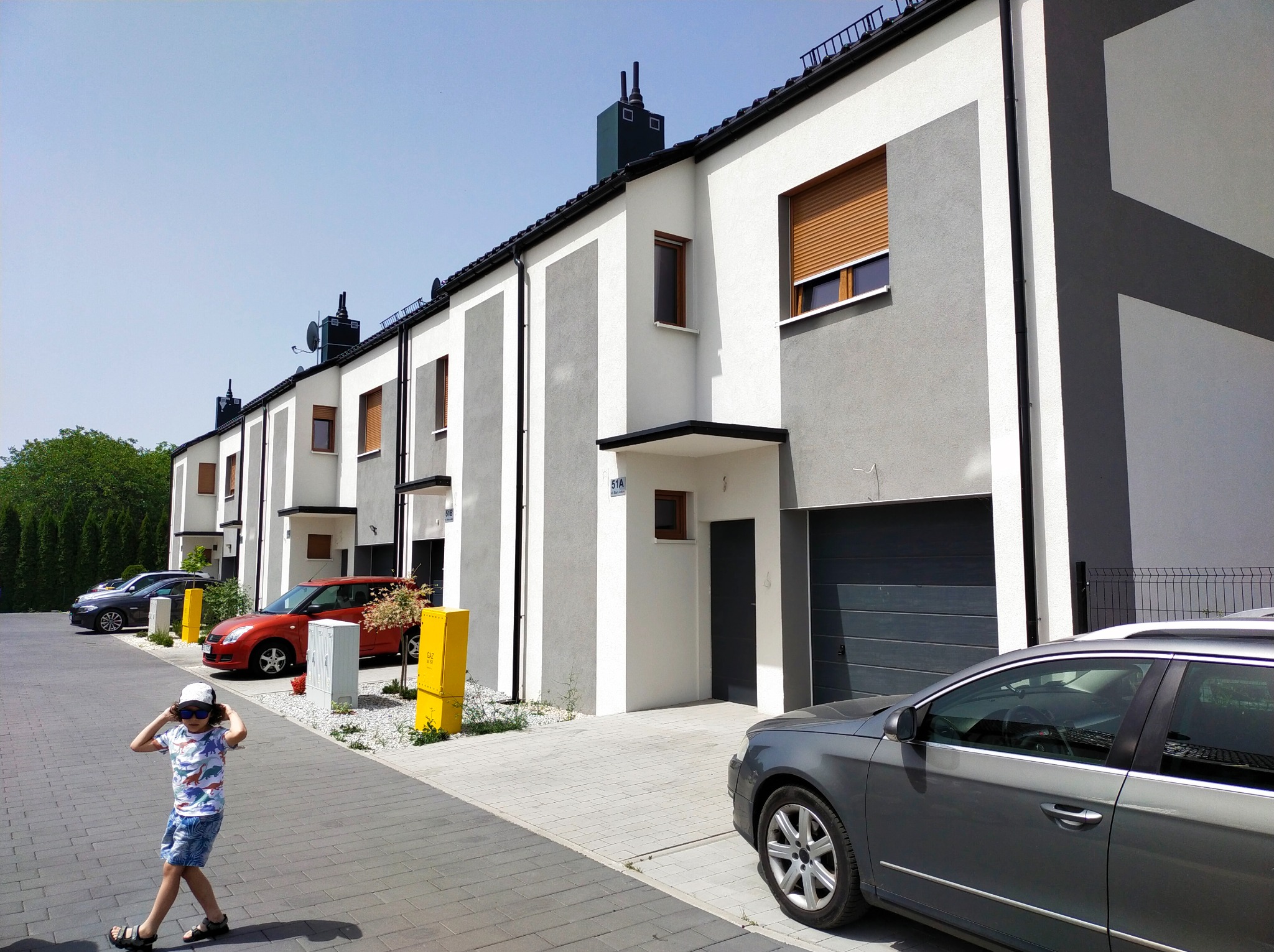 Szereg nowoczesnych domów jednorodzinnych z garażami, pomalowanych w biało-szare pasy, z zaparkowanymi samochodami i dzieckiem na chodniku.
