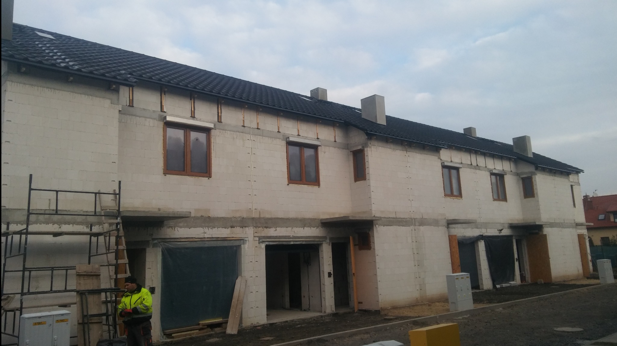 Budowa szeregowca z białego bloczka, widoczne okna z brązowymi ramami, czarny dach, rusztowanie przy ścianie i pracownik w jaskrawej kurtce.