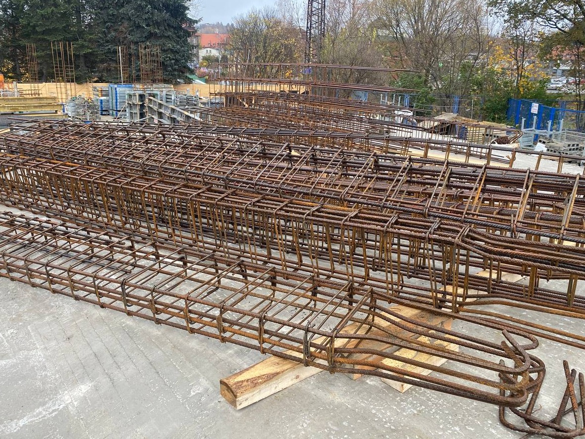 Siatka zbrojeniowa ułożona na placu budowy, widoczne elementy konstrukcyjne i drewniane podkładki, tło z drzewami i budynkami.