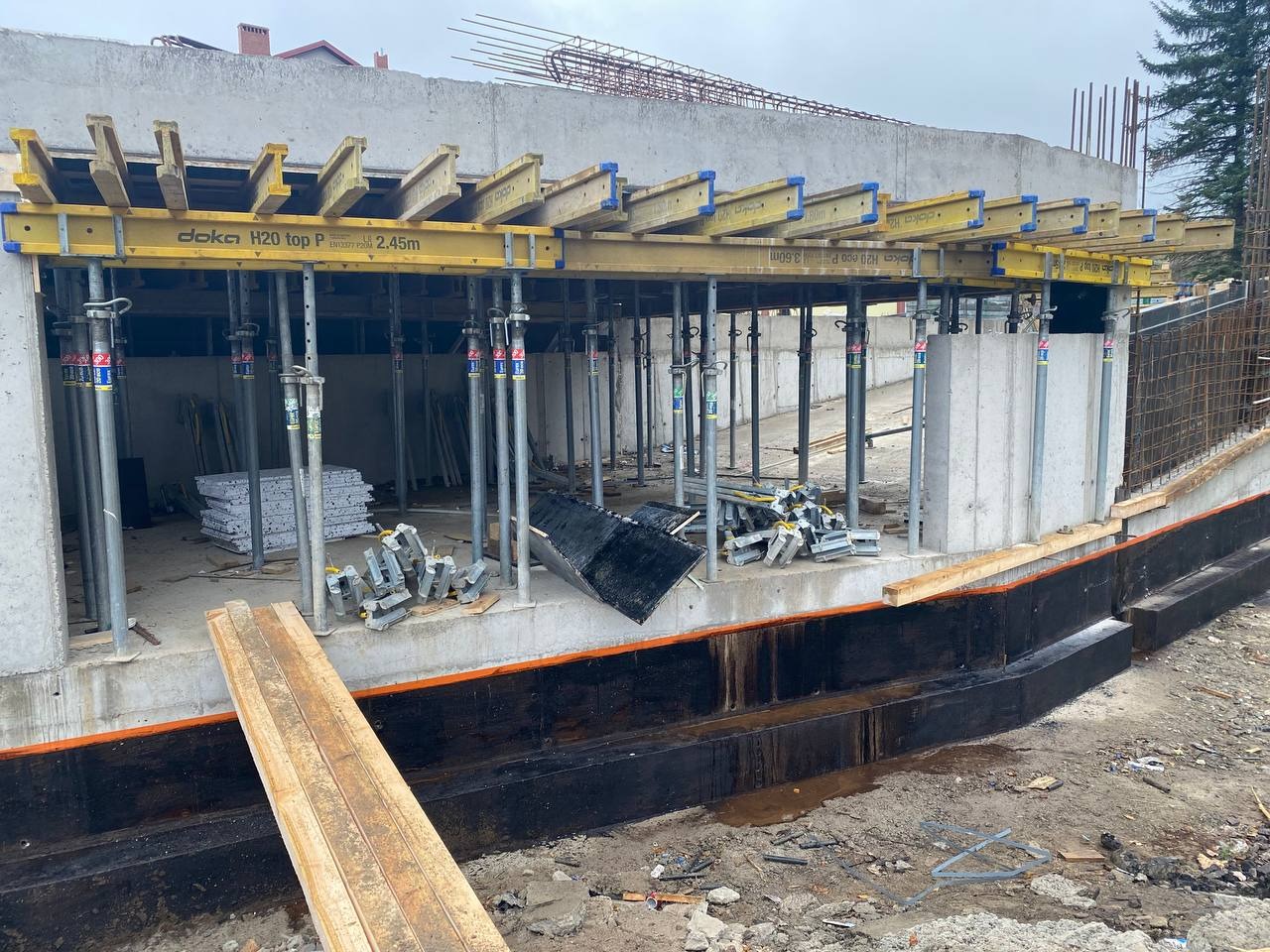 Wznoszenie fundamentów: Szalunek Doka H20 top P 2.45m podtrzymywany stalowymi stemplami, widoczne zbrojenie i izolacja przeciwwilgociowa, teren budowy w trakcie prac.