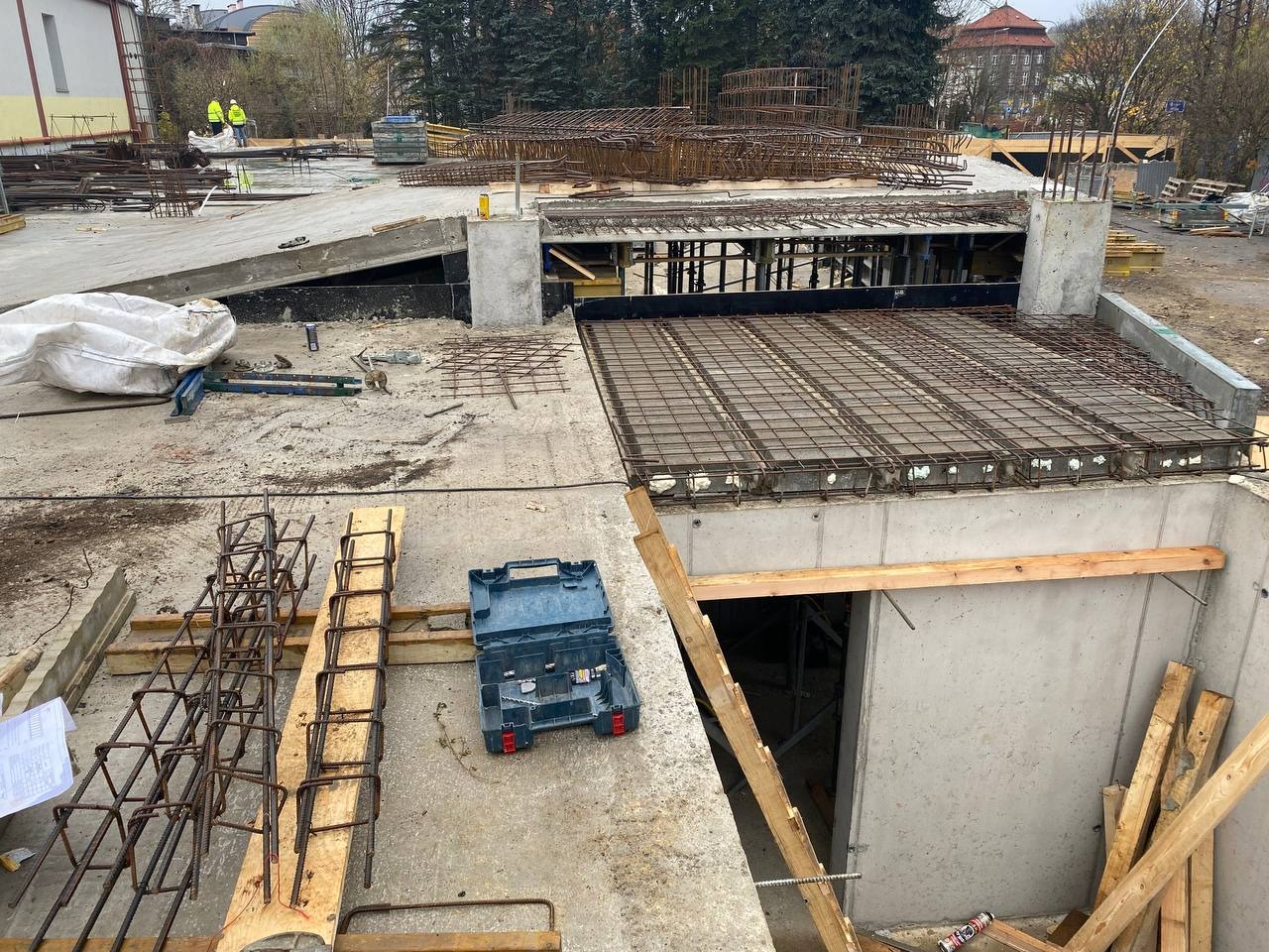 Widok z góry na plac budowy z częściowo zabetonowanym fundamentem, widoczne zbrojenia, szalunki i otwarte narzędzia w niebieskiej skrzynce, w tle budynek i drzewa.