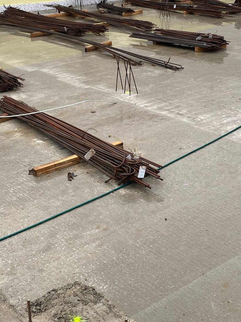 Wiązki prętów zbrojeniowych leżące na wilgotnej, betonowej płycie fundamentowej, oddzielone drewnianymi belkami, zielony wąż widoczny w tle.