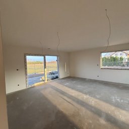 Dobra Robota Budowlana - Jasne, puste wnętrze po gładziowaniu ścian i wylewce podłogi, z widocznymi przewodami elektrycznymi zwisającymi z sufitu i dużym oknem z widokiem na zewnątrz.