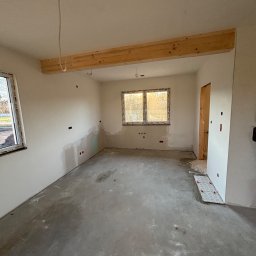 Dobra Robota Budowlana - Surowe wnętrze w trakcie prac wykończeniowych: ściany po gładzi, surowa podłoga, drewniana belka stropowa, okno z widokiem na zewnątrz. Widoczne punkty elektryczne.