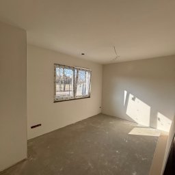 Dobra Robota Budowlana - Jasne, puste pomieszczenie po gładziowaniu ścian i wylewce podłogi, z oknem na zewnątrz. Widoczne gniazdko elektryczne i wystający kabel z sufitu. Świeże, minimalistyczne wnętrze.