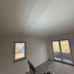 Dobra Robota Budowlana - Wnętrze w trakcie prac wykończeniowych: gładkie, białe ściany, okna z widokiem na zewnątrz i szlifierka do gładzi na pierwszym planie. Widoczne kable wystające z sufitu.