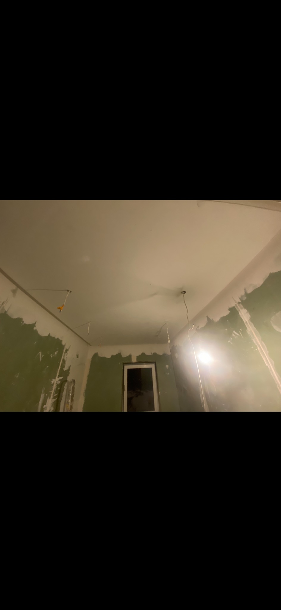 Pokój w trakcie remontu: zielone ściany z widocznymi śladami szpachlowania, sufit przygotowany do malowania, widoczne kable oświetleniowe i okno w tle.