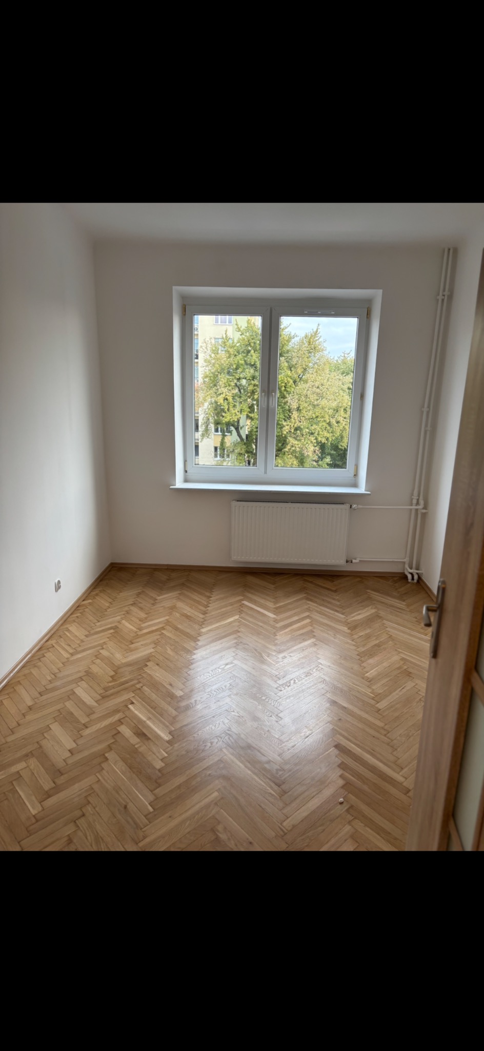 Jasne, odnowione wnętrze pokoju z parkietem ułożonym w jodełkę, białymi ścianami i oknem z widokiem na zieleń. Widoczny grzejnik pod oknem i fragment otwartych drzwi.