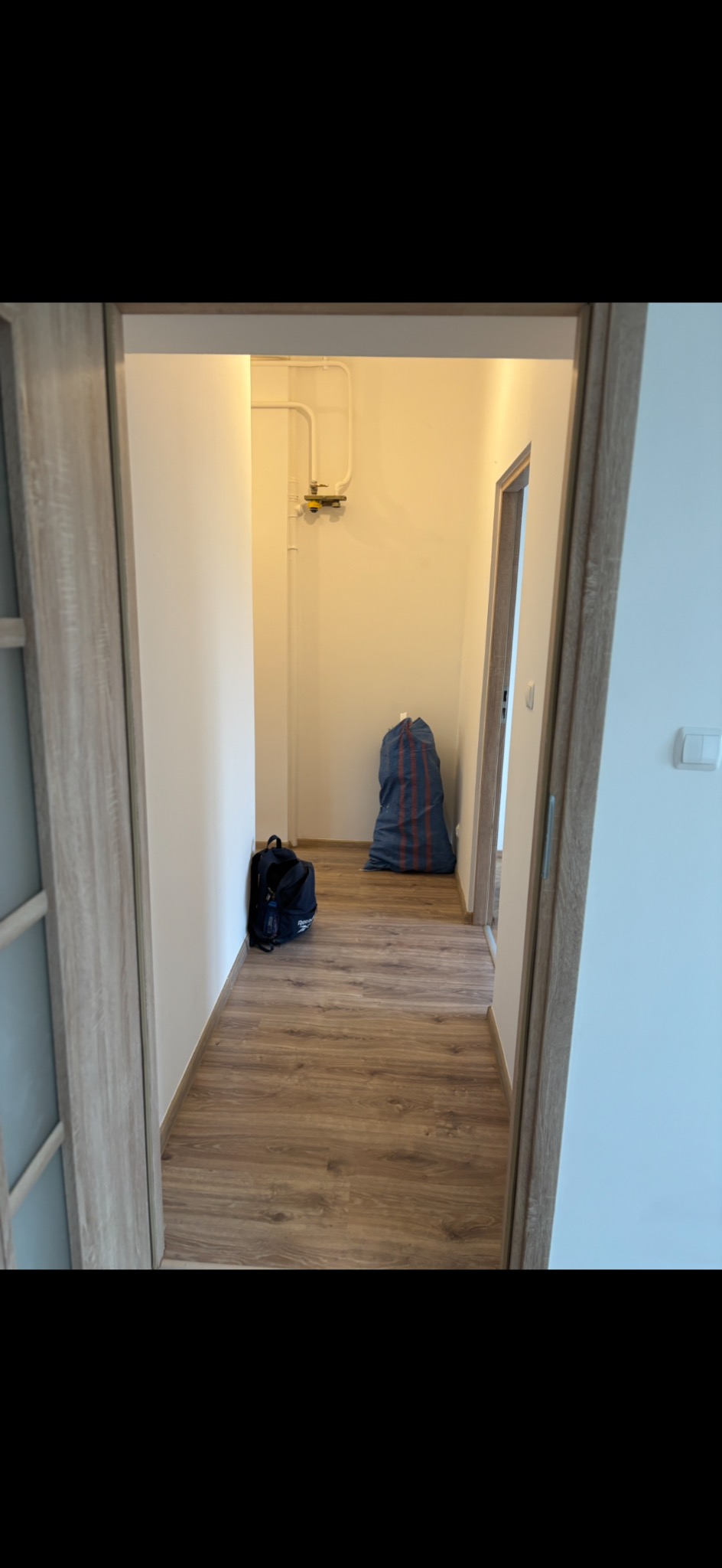 Korytarz po remoncie z nową podłogą drewnopodobną. Widoczne drzwi z ościeżnicą w kolorze drewna, torba i worek budowlany. Białe ściany i rury gazowe.