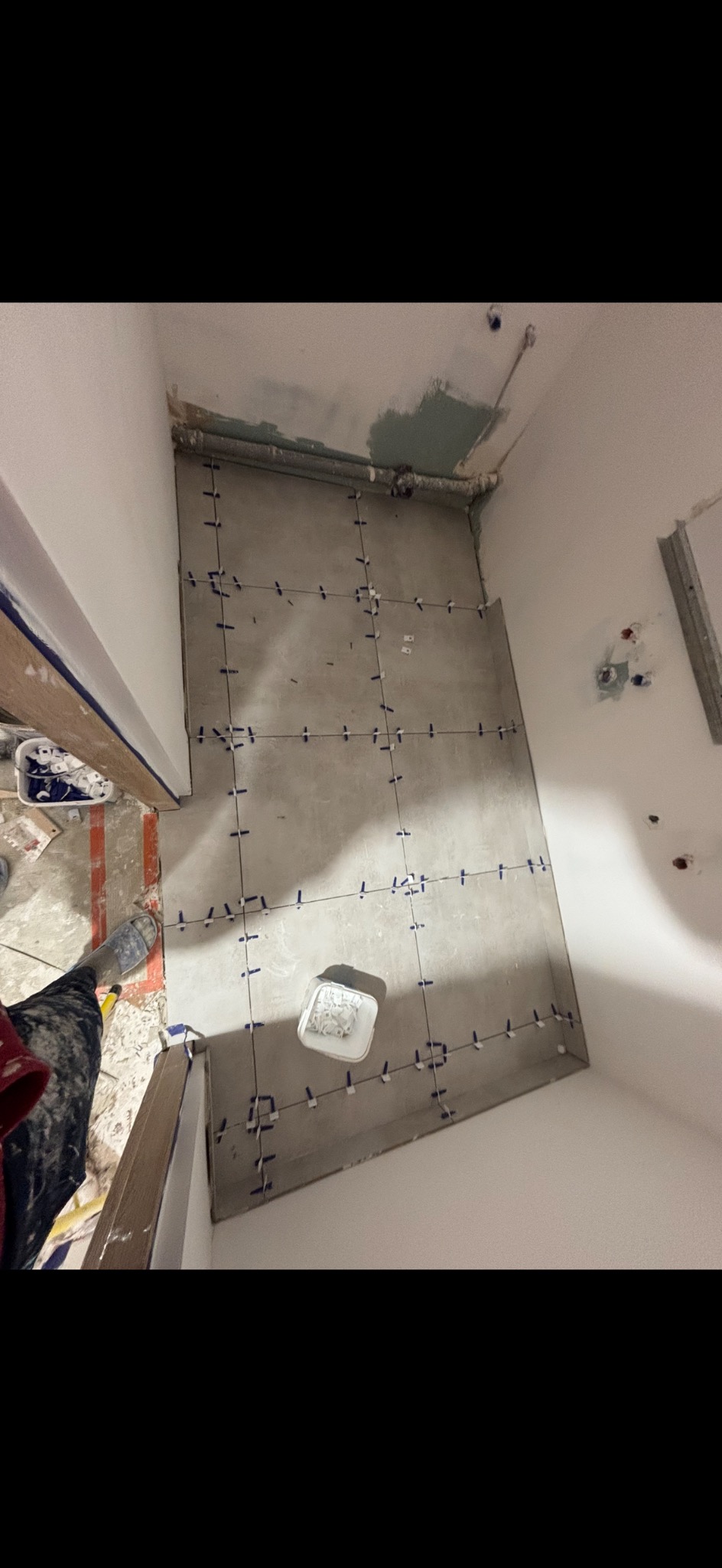 Szare płytki podłogowe w trakcie układania, z użyciem niebieskich krzyżyków dystansowych. Widoczny fragment ściany i podłogi w trakcie prac wykończeniowych.