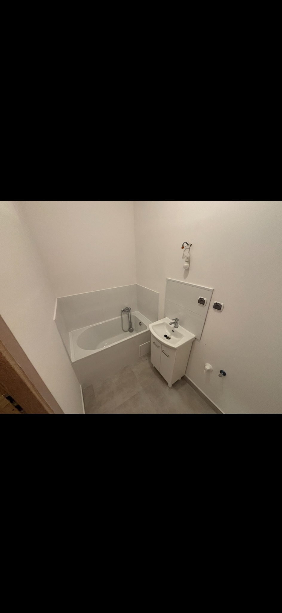 Biała, wykończona łazienka z wanną i umywalką z szafką. Widoczne szare płytki podłogowe i białe ściany. Minimalistyczny styl, świeżo po remoncie.