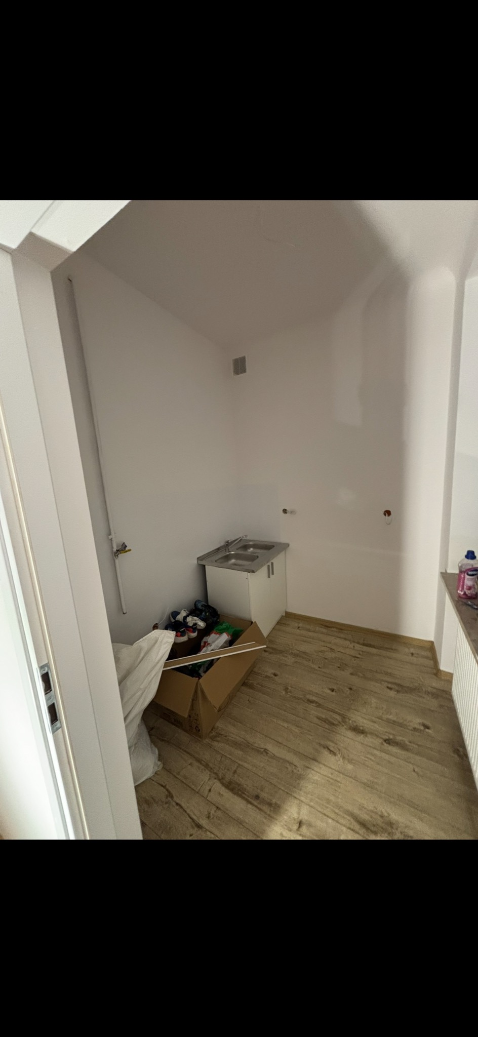 Wnętrze małej, białej łazienki z drewnopodobną podłogą. Widoczna biała szafka z umywalką, karton z rzeczami i rury przy ścianie. Minimalistyczny styl.