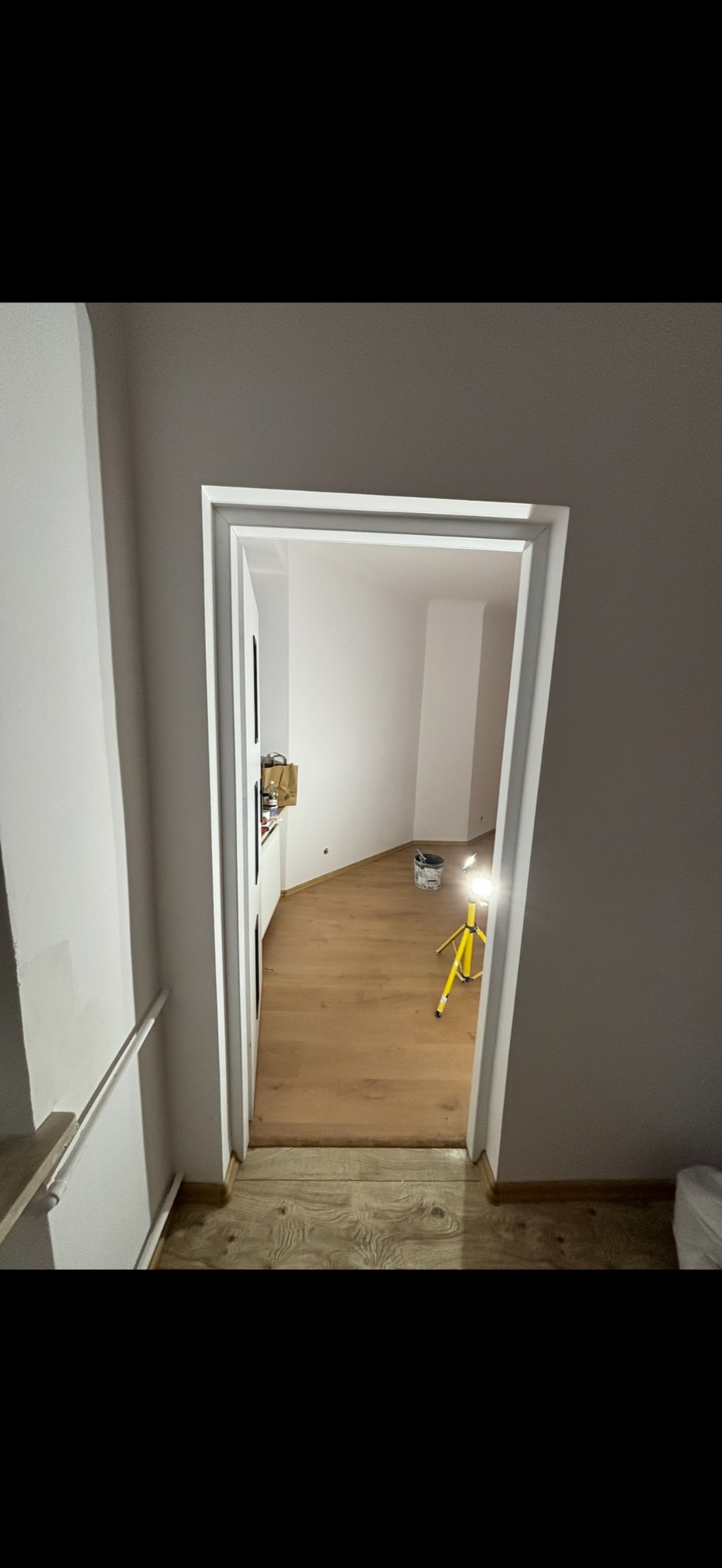 Białe, odnowione futryny drzwiowe w jasnym wnętrzu. Widoczna podłoga z paneli, ściany pomalowane na biało, w głębi pokoju wiadro z farbą i lampa na statywie.