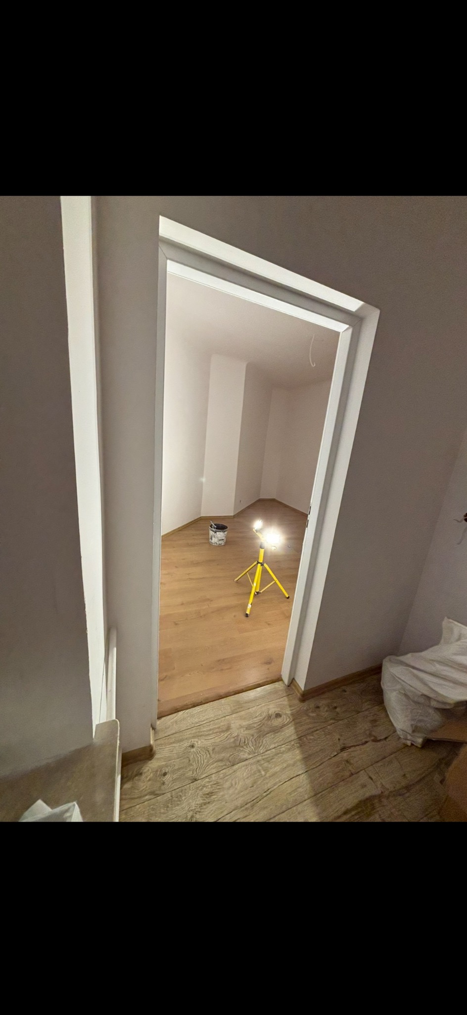 Wnętrze po odświeżeniu: białe ściany, drewniana podłoga, widoczna lampa budowlana i wiadro z farbą. Ujęcie przez futrynę drzwi.