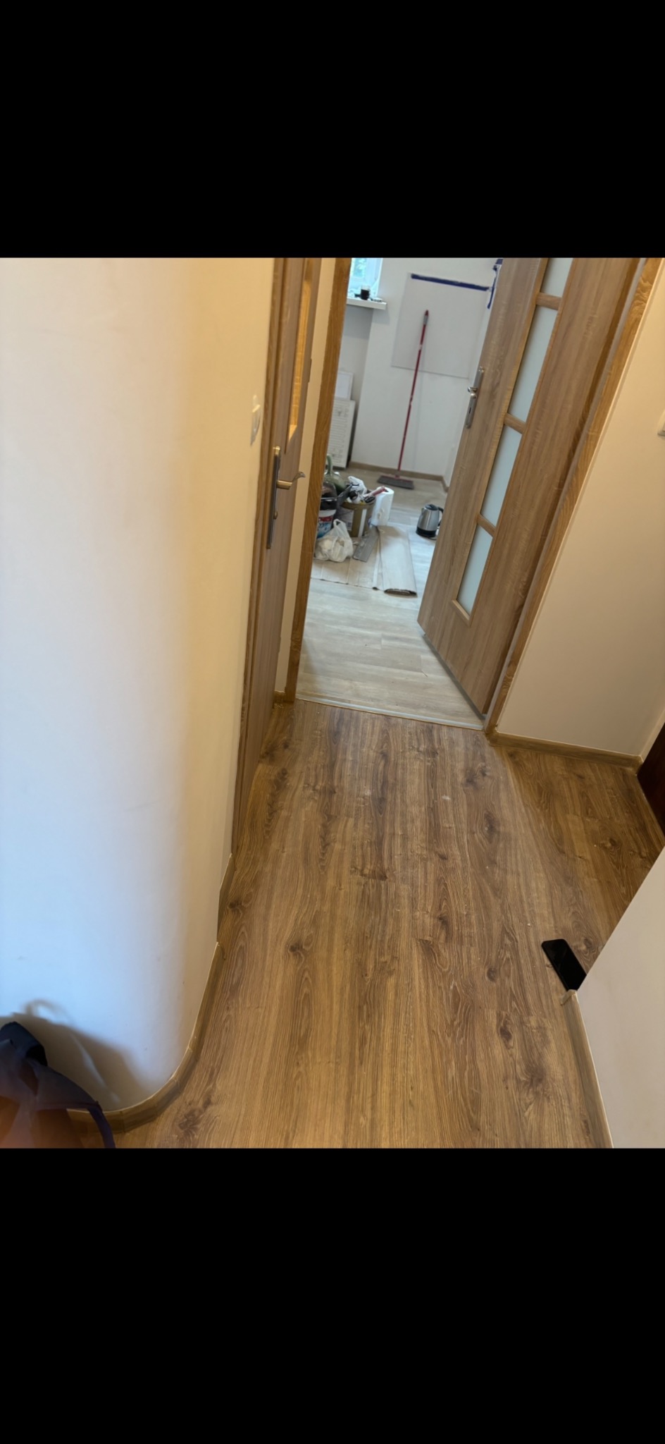 Ułożona podłoga z paneli drewnopodobnych w korytarzu, widoczne otwarte drzwi z przeszkleniami i fragment pomalowanej ściany. W tle widoczne narzędzia i materiały budowlane.