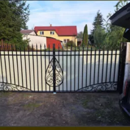 Nowoczesna brama wjazdowa z czarnymi, metalowymi prętami i dekoracyjnymi elementami, wypełniona mlecznym szkłem lub tworzywem, na tle domu z czerwoną dachówką i otwartym bagażnikiem samochodu.