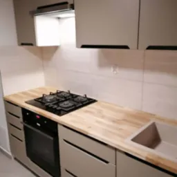 Jasna kuchnia z drewnianym blatem, czarną płytą grzewczą i minimalistycznymi uchwytami szafek.