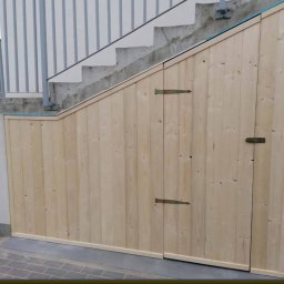 Marek - Drewniana zabudowa pod schodami zewnętrznymi z dwiema małymi furtkami, widoczne metalowe zawiasy i fragment betonowych schodów z metalową balustradą.