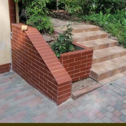 Marek - Zewnętrzne schody z jasnych płytek prowadzące do ogrodu, z ceglaną ścianką oporową i donicą, widok z dołu.