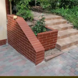 Zewnętrzne schody z jasnych płytek prowadzące do ogrodu, z ceglaną ścianką oporową i donicą, widok z dołu.