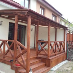 Marek - Zadaszony, drewniany taras z balustradą w geometryczny wzór, przylegający do domu, widoczne schody wejściowe i osoba w tle.