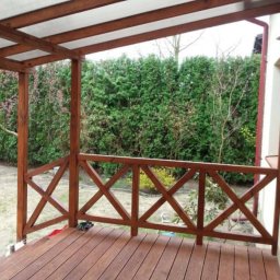 Marek - Zadaszony taras z drewnianą podłogą i balustradą w kształcie krzyży, widok na zielony ogród z tujami.