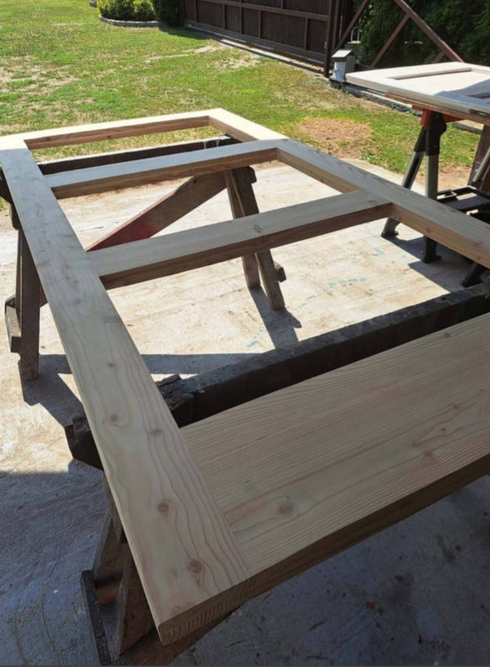 Drewniana rama konstrukcyjna, być może fragment większej konstrukcji, ustawiona na kozłach stolarskich na zewnątrz, w tle trawnik i ogrodzenie.