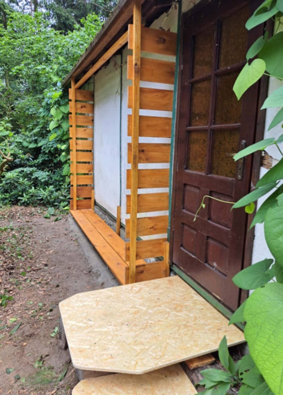 Surowa konstrukcja drewniana przy wejściu do budynku z brązowymi drzwiami, widoczne deski i płyty OSB, otoczenie w zieleni.