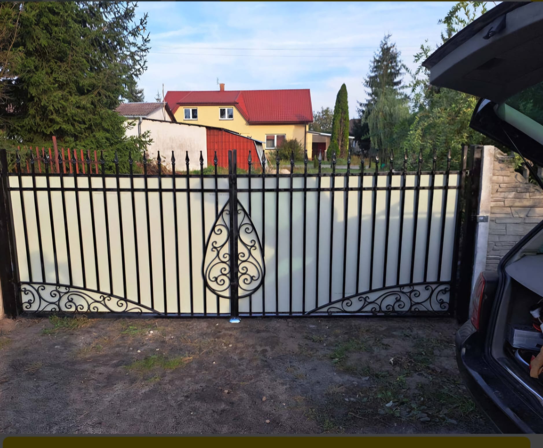 Nowoczesna brama wjazdowa z czarnymi, metalowymi prętami i dekoracyjnymi elementami, wypełniona mlecznym szkłem lub tworzywem, na tle domu z czerwoną dachówką i otwartym bagażnikiem samochodu.