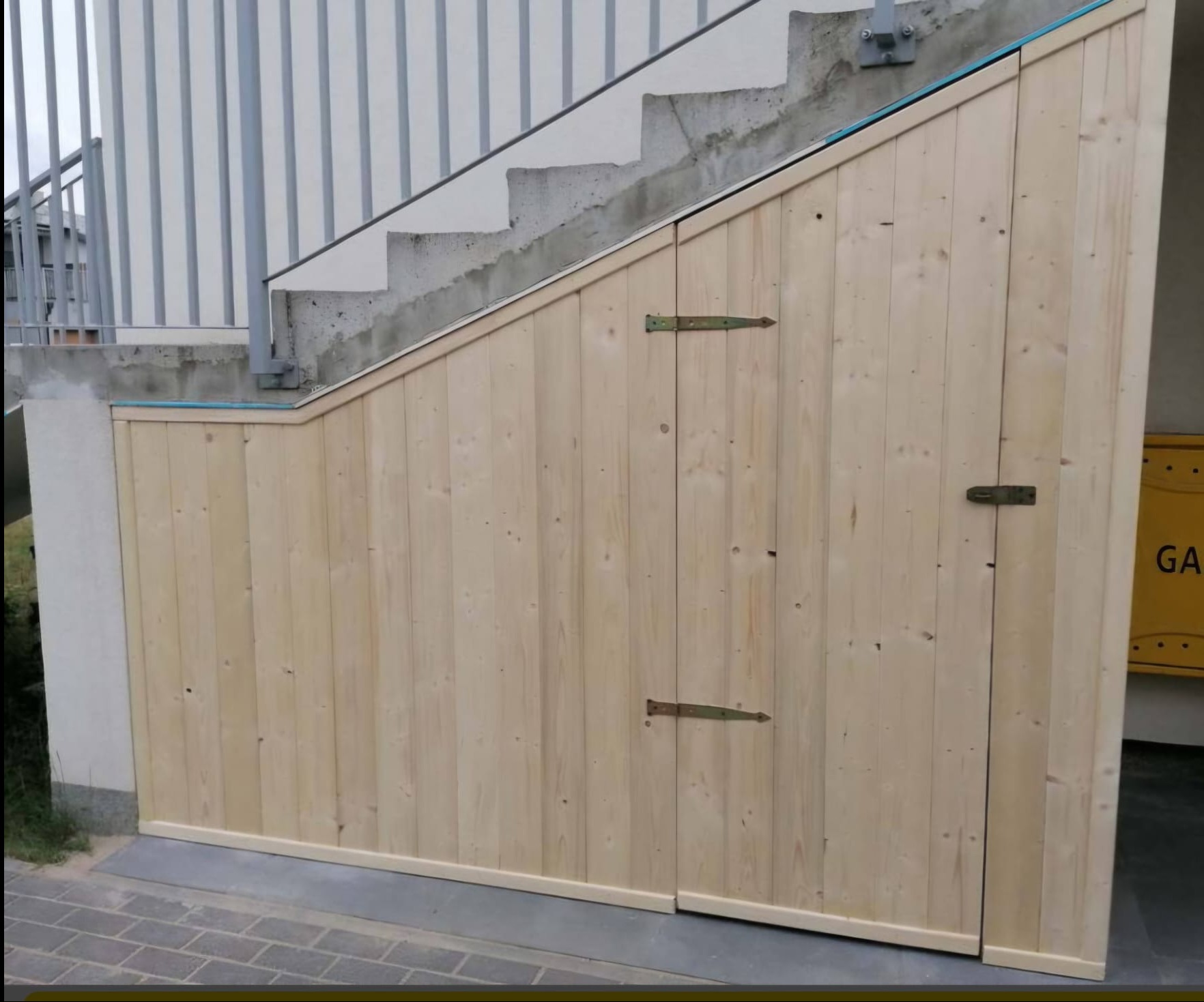 Drewniana zabudowa pod schodami zewnętrznymi z dwiema małymi furtkami, widoczne metalowe zawiasy i fragment betonowych schodów z metalową balustradą.