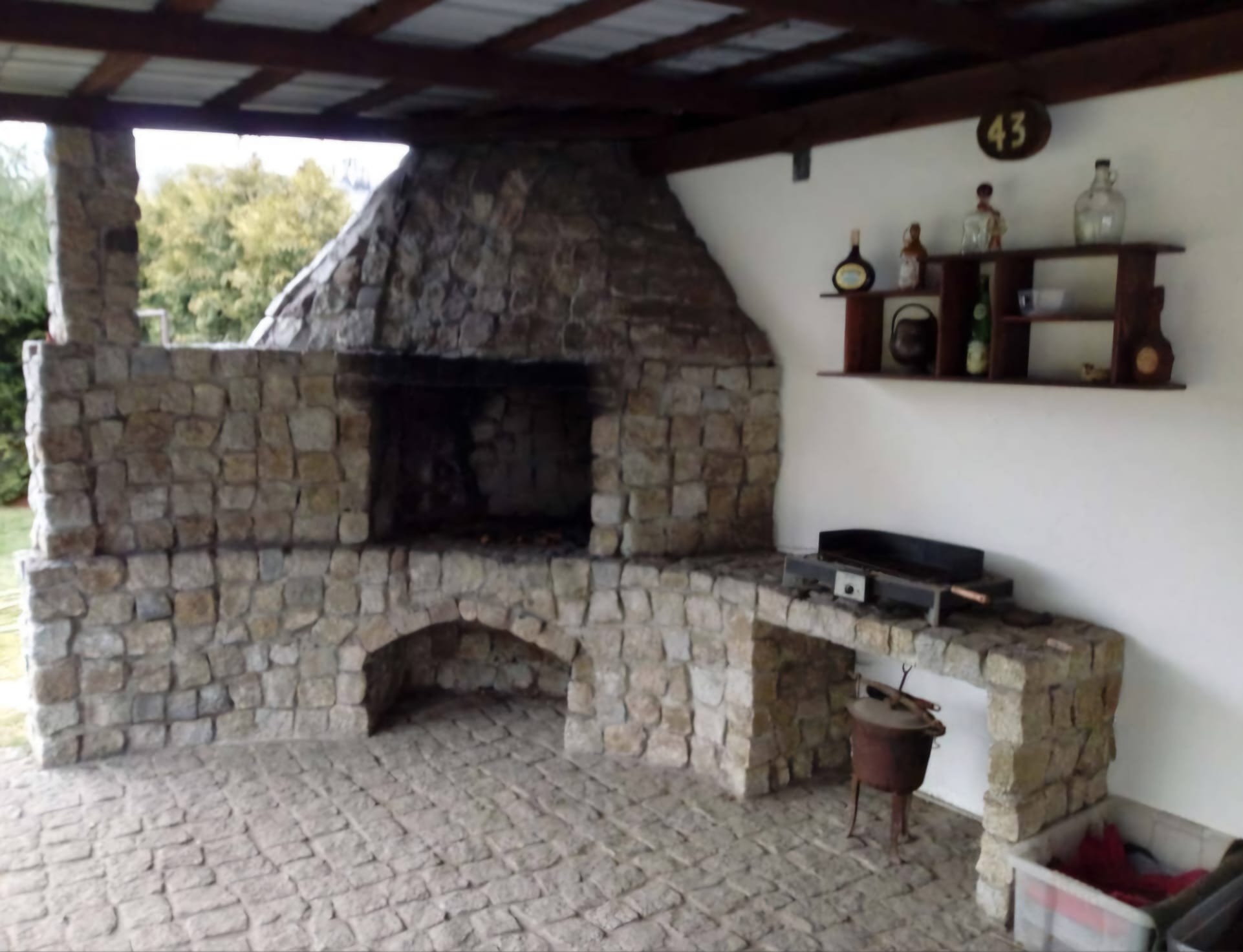 Kamienny grill ogrodowy z wędzarnią pod zadaszeniem, z blatem roboczym i półką na akcesoria, numer domu 43 na ścianie.