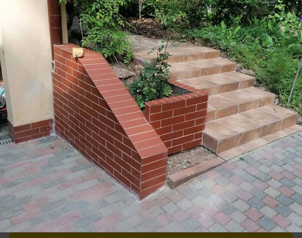 Zewnętrzne schody z jasnych płytek prowadzące do ogrodu, z ceglaną ścianką oporową i donicą, widok z dołu.