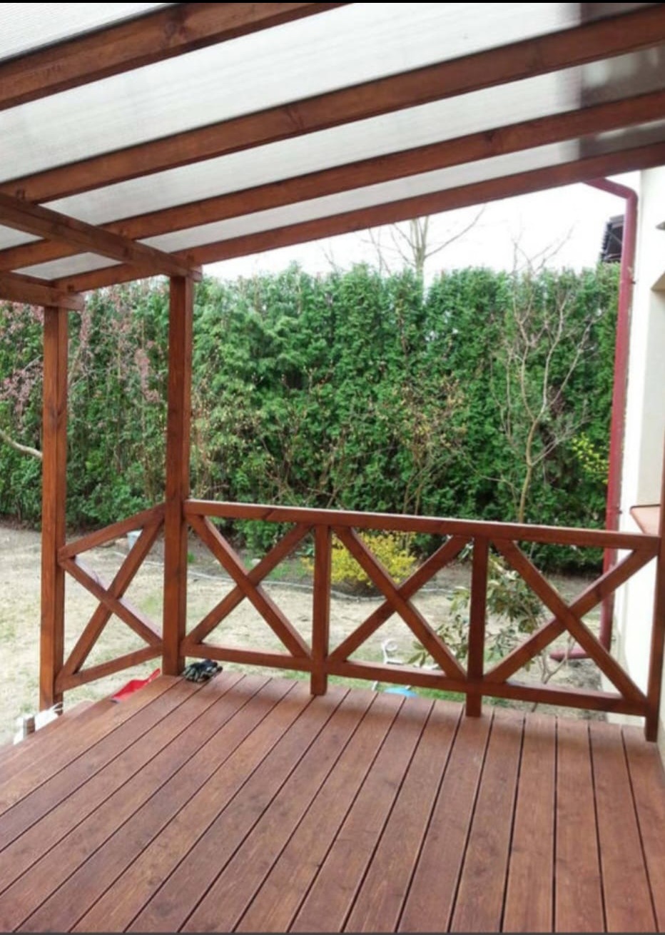 Zadaszony taras z drewnianą podłogą i balustradą w kształcie krzyży, widok na zielony ogród z tujami.