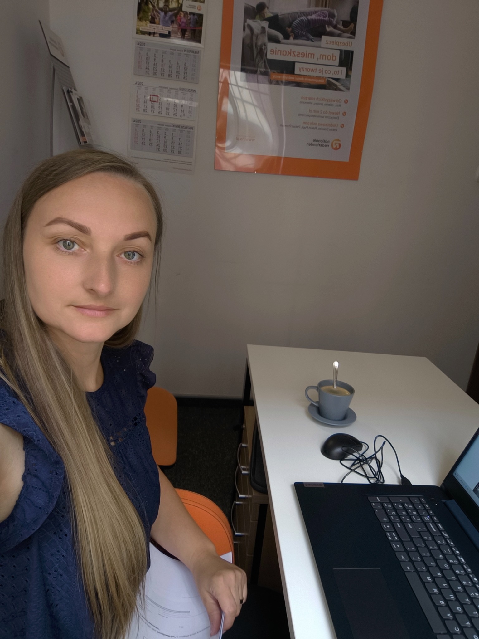 Kobieta z długimi blond włosami siedzi przy biurku z laptopem, myszką i filiżanką kawy. Na ścianie wisi kalendarz i plakat z ofertą ubezpieczeniową.