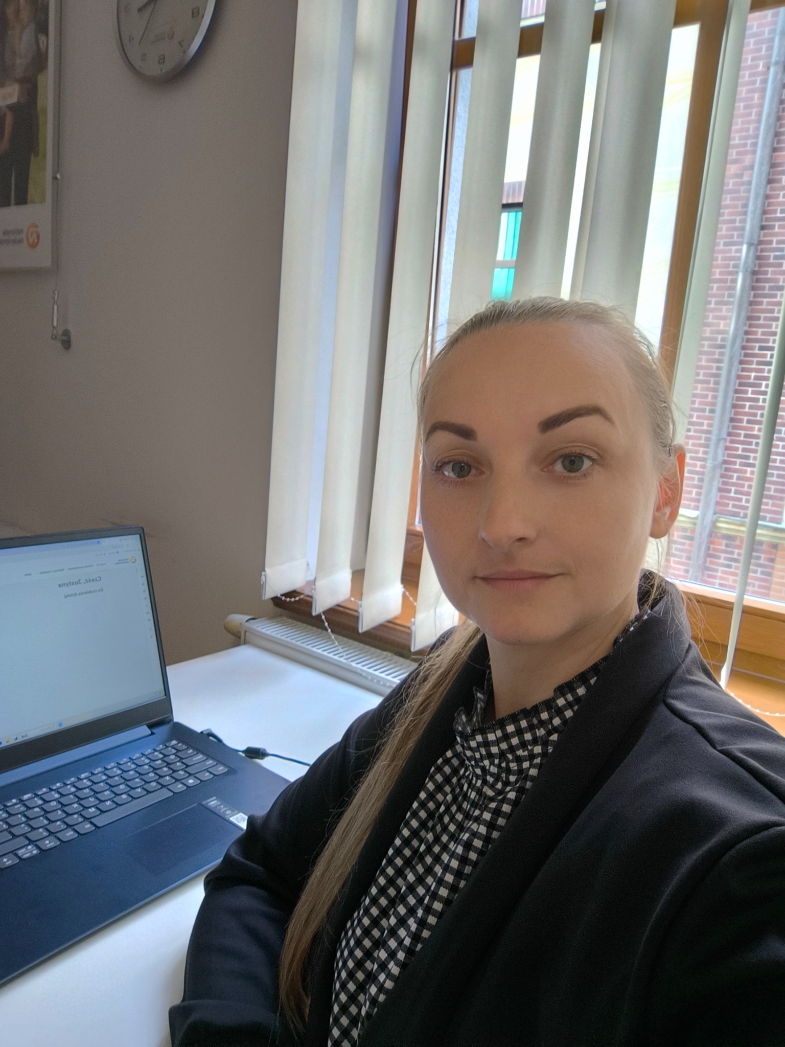 Portret kobiety w biurze przed laptopem, w tle okno z żaluzjami i ceglana ściana budynku za oknem, na ścianie w tle plakat.