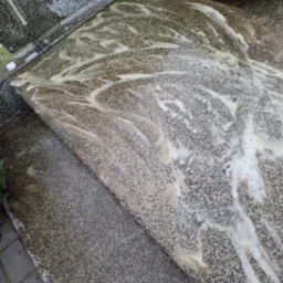 Granitowy nagrobek pokryty pianą czyszczącą, widoczne ślady spływającego detergentu, obok kostka brukowa i fragment krzewu.