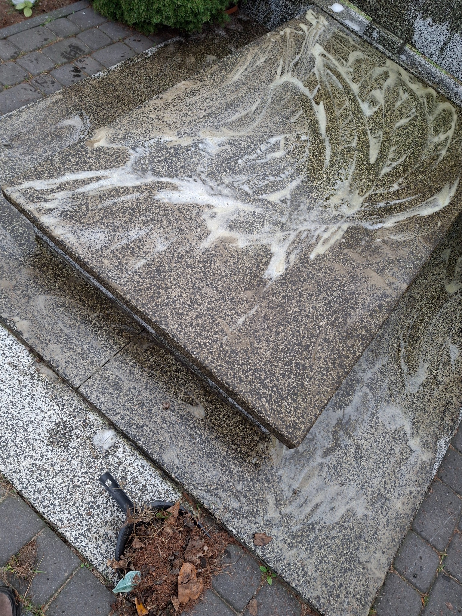 Czyszczenie nagrobka z szarego kamienia, widoczne ślady po myciu i resztki liści w czarnej szufelce obok.