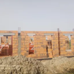 Wznoszone ściany z pustaków ceramicznych z widocznymi otworami okiennymi i zbrojeniami pionowymi, na pierwszym planie hałda ziemi.