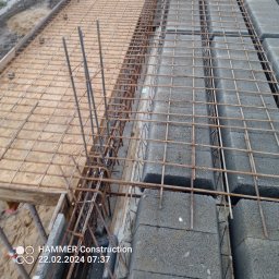 BULID - Zbrojenie i bloczki betonowe przygotowane do wylewki fundamentów, widoczne drewniane szalunki i wystające pręty zbrojeniowe.