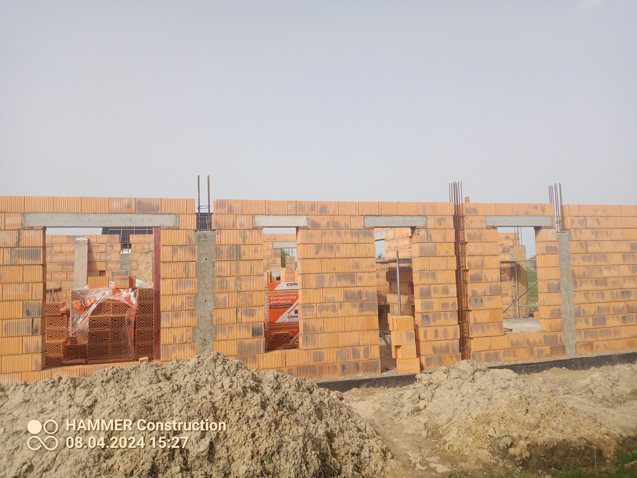 Wznoszone ściany z pustaków ceramicznych z widocznymi otworami okiennymi i zbrojeniami pionowymi, na pierwszym planie hałda ziemi.