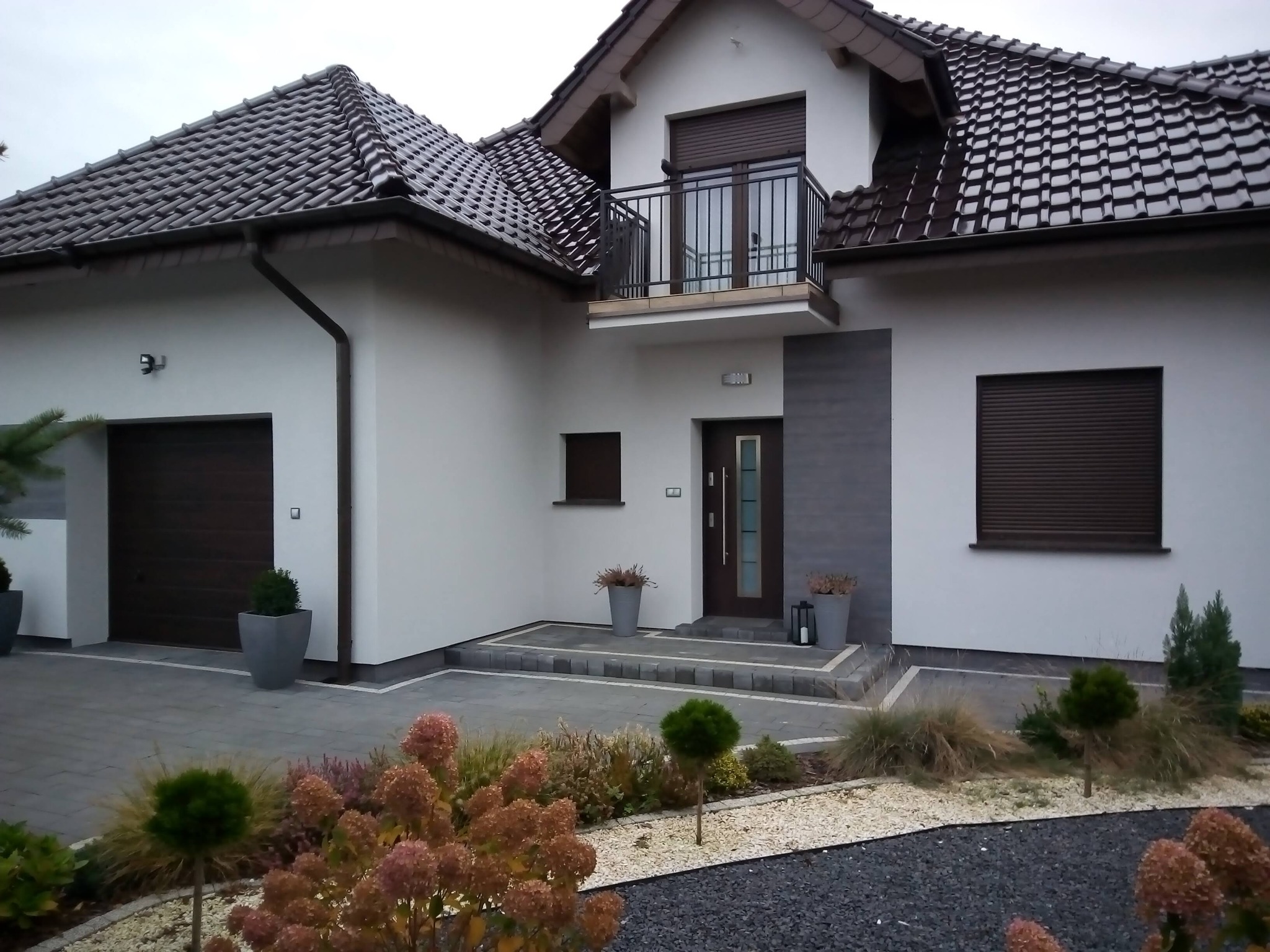 Nowoczesny dom jednorodzinny z brązową stolarką okienną, balkonem i zadbanym ogrodem z kamieniami i roślinami ozdobnymi.