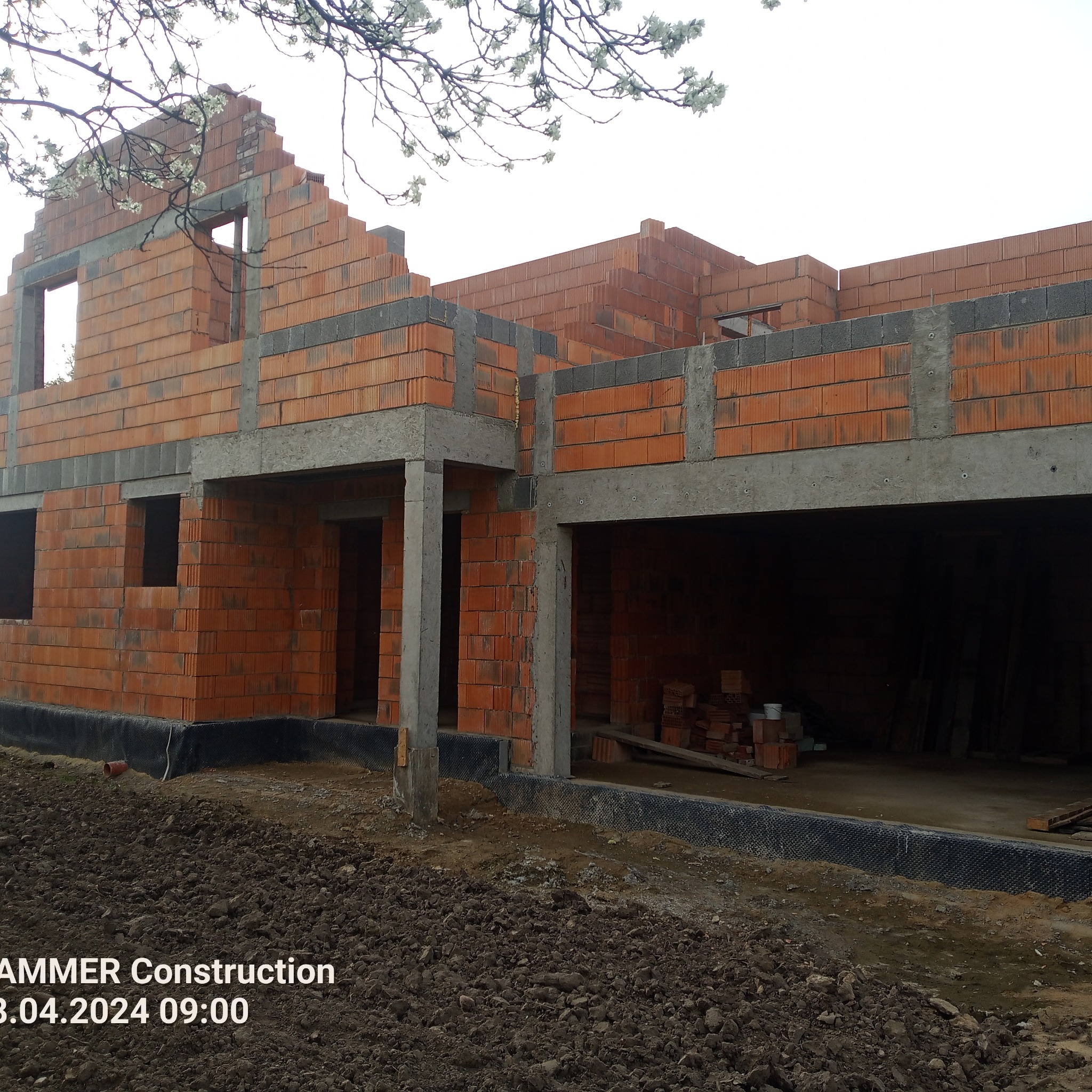 Surowy stan budynku w trakcie murowania ścian zewnętrznych z czerwonej cegły ceramicznej, widoczne betonowe nadproża i słupy, z zewnątrz ziemia i kwitnące drzewo w tle.