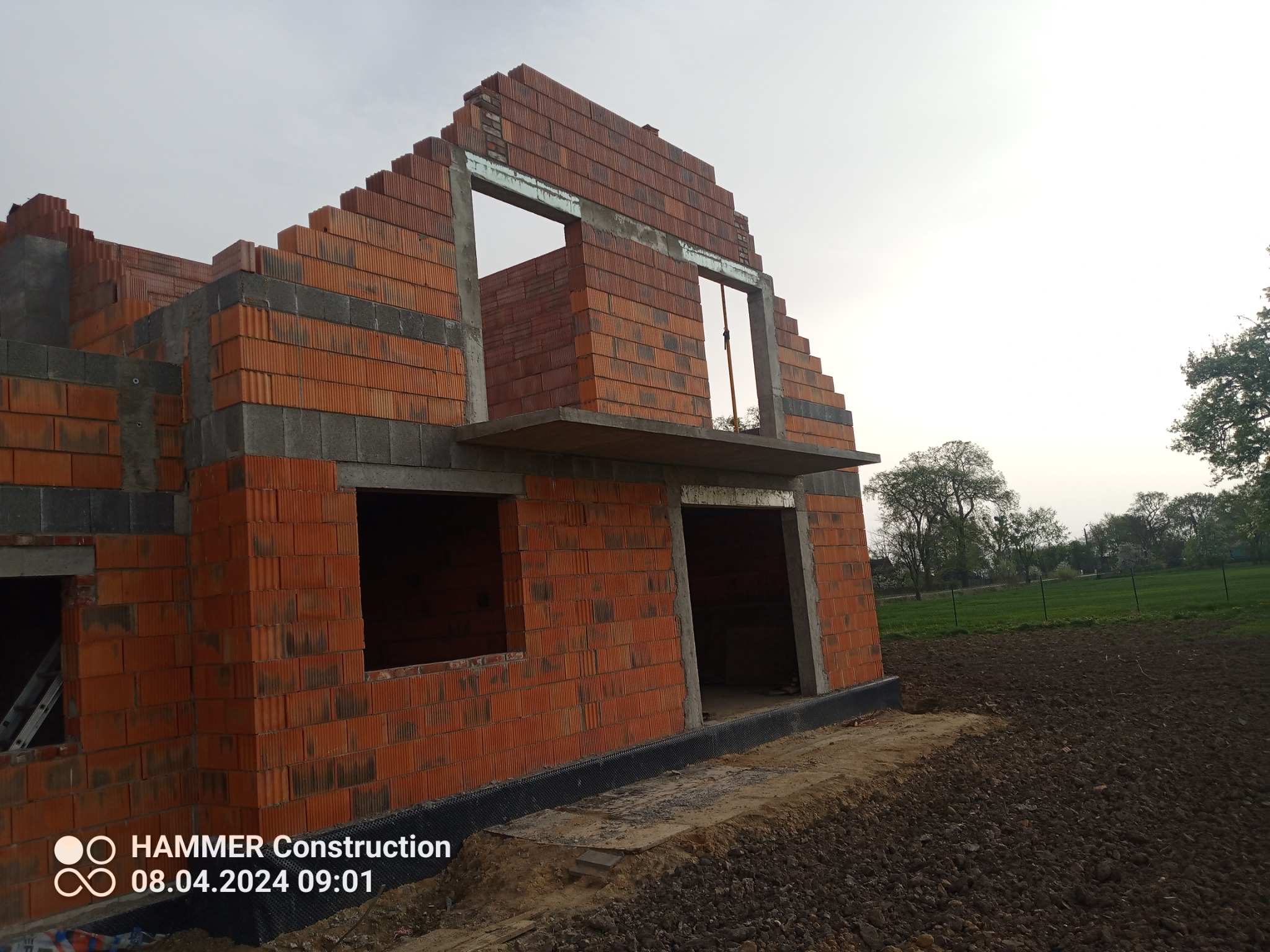 Surowy stan budynku jednorodzinnego w trakcie murowania ścian z czerwonej cegły ceramicznej, widoczne otwory okienne i drzwiowe, zarys balkonu, na pierwszym planie przygotowany teren pod budowę.